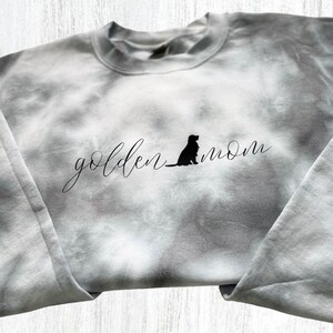 Pu&ograve; includere: Una felpa grigia tie-dye con il testo "golden mom" e una silhouette di un cane in nero.