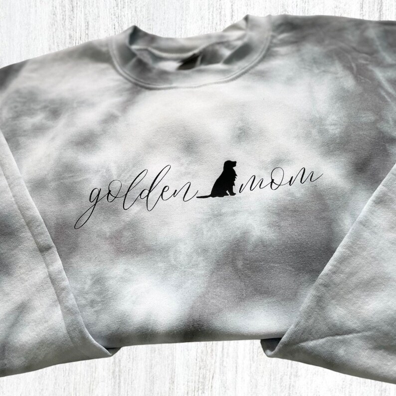 Pu&ograve; includere: Una felpa grigia tie-dye con il testo "golden mom" e una silhouette di un cane in nero.