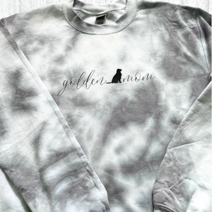 Pu&ograve; includere: Una felpa color celeste tie-dye con il testo "golden mom" e la silhouette di un cane in nero.