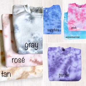 Pu&ograve; includere: Un mucchio di felpe tie-dye in vari colori, tra cui beige, rosa, grigio, acquamarina, rosa, zaffiro e viola. Le felpe sono piegate e disposte in fila.