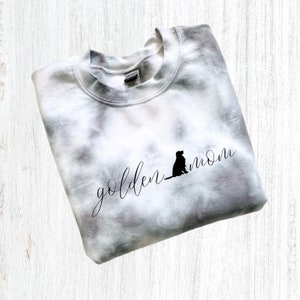 Pu&ograve; includere: Una felpa bianca tie-dye con il testo "golden mom" e la silhouette di un golden retriever.