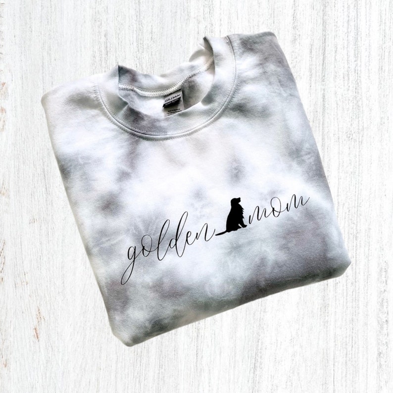 Pu&ograve; includere: Una felpa bianca tie-dye con il testo "golden mom" e la silhouette di un golden retriever.