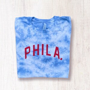May include: A blue tie-dye t-shirt with the word "PHILA." printed in red on the front.