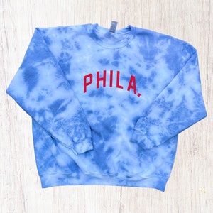 May include: A blue tie-dye sweatshirt with the word "PHILA." embroidered in red on the front.