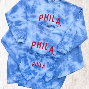May include: Three blue tie-dye sweatshirts with the word "PHILA." embroidered in red on each. The sweatshirts are layered on top of each other, with the smallest sweatshirt on top.