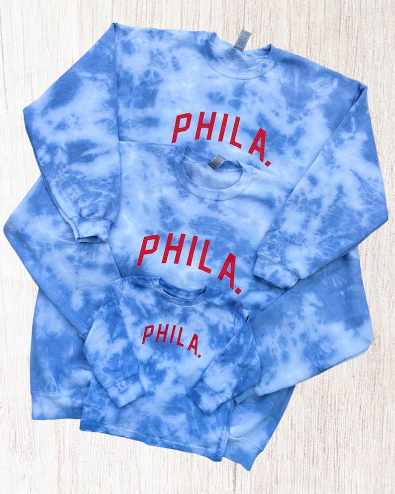May include: Three blue tie-dye sweatshirts with the word "PHILA." embroidered in red on each. The sweatshirts are layered on top of each other, with the smallest sweatshirt on top.