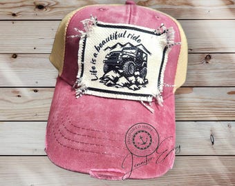 May include: A red and beige distressed baseball cap with a patch that says "Life is a beautiful ride" and features a black jeep driving on a dirt road. The cap also has a small anchor logo with the text "Anchored Hope".