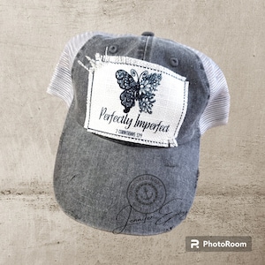 May include: Gray trucker hat with a white mesh back and a distressed look. The front patch features a butterfly design and the words "Perfectly Imperfect" with a Bible verse reference. The hat has a curved brim.