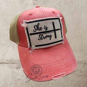 May include: A pink and beige trucker hat with a distressed look. The hat has a patch on the front that says "She is Strong" with a cross and the words "Proverbs 3:15".