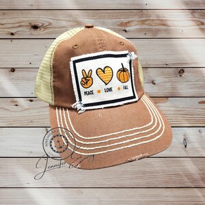 May include: Brown and tan trucker hat with a distressed brim. The hat features a patch with the words "Peace Love Fall" and corresponding symbols. White stitching accents the brim.