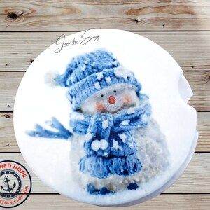 May include: A white ceramic coaster with a blue and white snowman wearing a blue knitted hat and scarf. The coaster has the text "Anchored Hope Christian Clothing Inc." in a red circle with a white anchor.