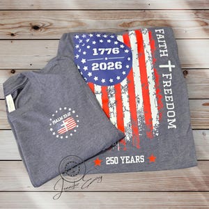 May include: Two gray t-shirts with patriotic designs. One features a distressed American flag with "1776 2026" and "Faith Freedom" text. The other has a smaller flag design and "Psalm 33:12". Both shirts are folded on a wooden surface.