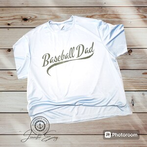 May include: A light blue t-shirt with the text "Baseball Dad" in a green script font. The shirt is folded and laid on a wooden surface. The text "Anchored Hope Christian Clothing" is visible in a small circle on the bottom of the shirt.