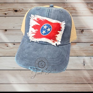 May include: A distressed denim baseball cap with a tan mesh back. The front features a red, white, and blue patch of the Tennessee state flag. The cap has a pre-curved bill and a distressed, worn look.