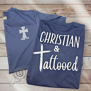 May include: Two blue t-shirts. One features a white Celtic cross and the text "John 7:24". The other shirt says "CHRISTIAN & Tattooed" in white. Both shirts are folded and displayed on a light wood grain surface.