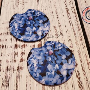 May include: Two car coasters with a floral design. The coasters feature a pattern of blue and white hydrangea flowers against a dark background. The coasters are round with a small notch on one side.