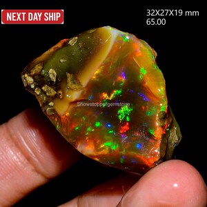 Könnte beinhalten: Ein roher Opal-Edelstein mit lebendigen Farben, darunter Rot, Grün und Blau, vor dunklem Hintergrund. Der Opal misst ca. 3,2 x 2,7 x 1,9 cm. Der Text "Showstoppergemstone" ist sichtbar.