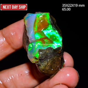 Könnte beinhalten: Ein roher Opal-Edelstein mit leuchtenden Farben, darunter Grün, Orange und Blau. Der Stein misst ca. 3,5 x 2,2 x 1,9 cm. Der Text "Next Day Ship" ist in der oberen linken Ecke sichtbar.