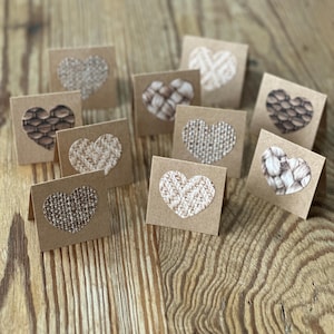 May include: A collection of small, square, kraft paper cards with heart-shaped cutouts. The cutouts reveal various textured patterns, including woven fabric and other designs. The cards are arranged on a wooden surface.