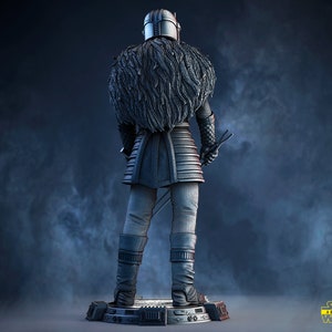 May include: A detailed, silver-colored figurine of a character from Star Wars wearing a fur-lined cloak and holding a lightsaber. The figurine is standing on a black base with the Star Wars logo.