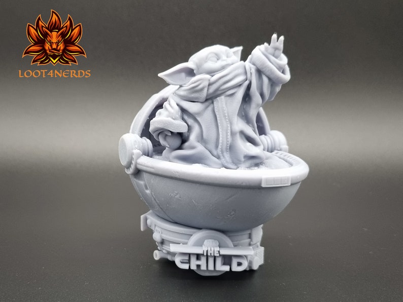 May include: A gray 3D printed figurine of a character from the Star Wars series, The Mandalorian. The character is sitting in a round pod with the text "The Child" on the base.