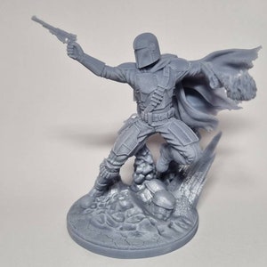 Puede incluir: Una figura gris de pl&aacute;stico impresa en 3D de un personaje mandaloriano de la franquicia Star Wars. El personaje sostiene un bl&aacute;ster en una mano y tiene una capa que fluye detr&aacute;s de &eacute;l. La figura est&aacute; de pie sobre una base rocosa.