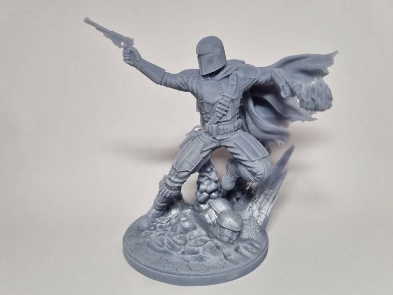 Puede incluir: Una figura gris de pl&aacute;stico impresa en 3D de un personaje mandaloriano de la franquicia Star Wars. El personaje sostiene un bl&aacute;ster en una mano y tiene una capa que fluye detr&aacute;s de &eacute;l. La figura est&aacute; de pie sobre una base rocosa.