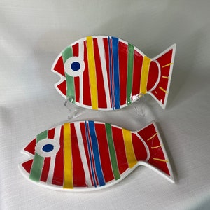May include: Two ceramic fish-shaped plates with red, yellow, blue, and green stripes. The plates are white with a glossy finish.