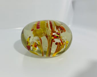 May include: A clear, oval-shaped glass paperweight with colorful floral designs in yellow, red, and orange. The interior features air bubbles, adding depth to the decorative piece. The paperweight is a decorative desk accessory.