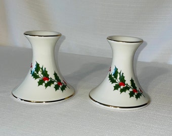 May include: Two white ceramic candle holders with a gold rim and a holly berry design. The candle holders have a flared base and a slender neck. The holly design features green leaves and red berries.