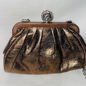 Pode incluir: Uma clutch bronze com fecho de bola prateada. A bolsa tem um design plissado e texturizado e uma al&ccedil;a de corrente prateada. O fecho &eacute; uma bola prateada decorativa com um intrincado padr&atilde;o de redemoinho.