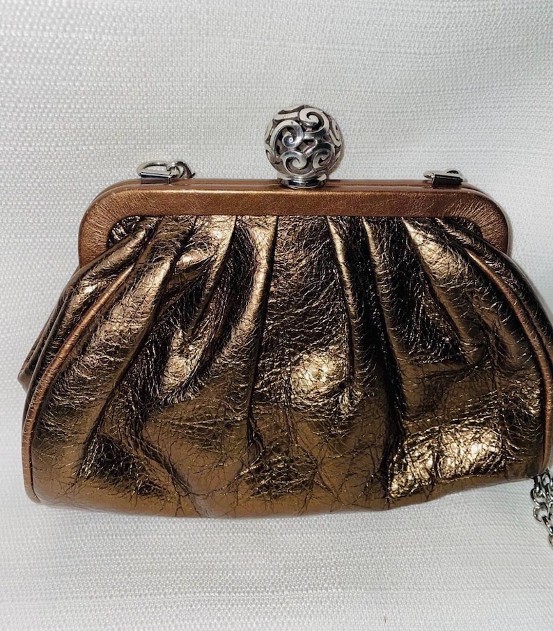 Pode incluir: Uma clutch bronze com fecho de bola prateada. A bolsa tem um design plissado e texturizado e uma al&ccedil;a de corrente prateada. O fecho &eacute; uma bola prateada decorativa com um intrincado padr&atilde;o de redemoinho.