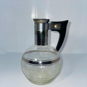May include: A clear glass pitcher with a round body and a silver band around the middle. It has a silver top and a black handle. The pitcher is likely for serving beverages.