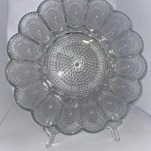 May include: Clear glass egg serving platter with a scalloped edge and individual egg compartments. The center features a textured, dotted design. The platter is supported by a clear plastic stand.