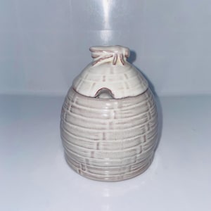 May include: A small, light beige ceramic honey pot with a matching lid. The pot has a textured, woven basket design. The lid has a small opening for a spoon. The honey pot is a kitchenware item.