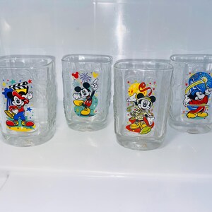 May include: Four clear, square-shaped drinking glasses with textured sides. Each glass features a different colorful image of Mickey Mouse in various poses. The glasses are displayed on a white surface.