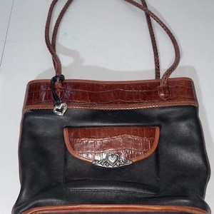 May include: Black and brown leather tote bag with a braided handle. The bag features a brown crocodile-textured leather trim and a front pocket with a silver heart-shaped embellishment. The bag has a structured design.