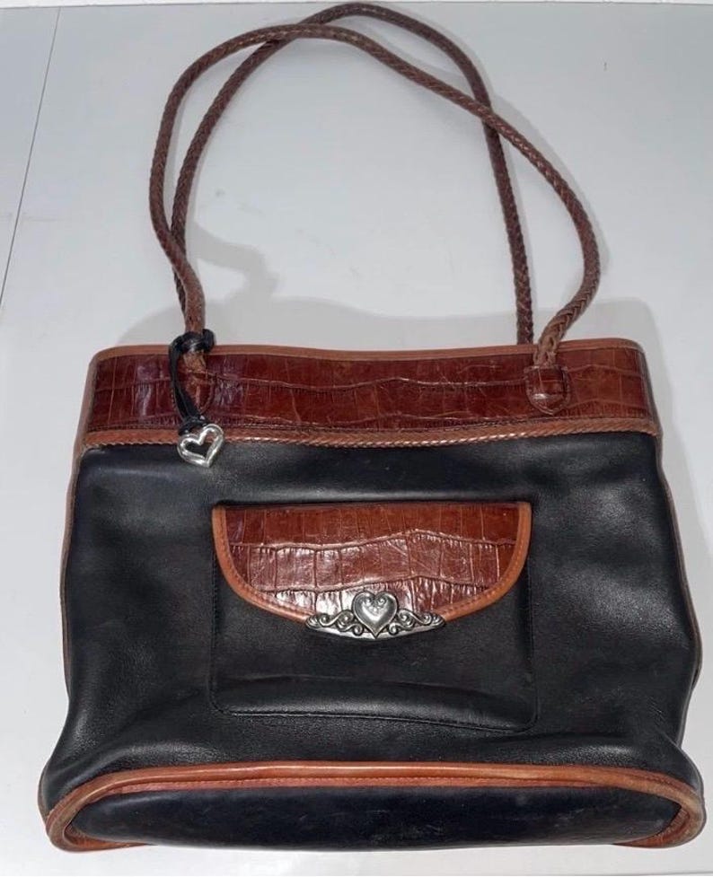 May include: Black and brown leather tote bag with a braided handle. The bag features a brown crocodile-textured leather trim and a front pocket with a silver heart-shaped embellishment. The bag has a structured design.