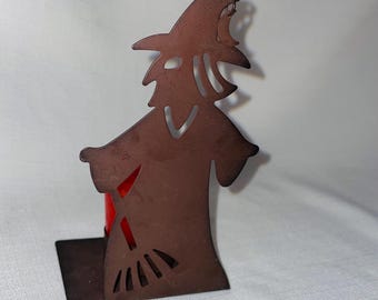 May include: A metal candle holder shaped like a witch holding a broom. The holder is brown and has a cutout design. A red candle is placed in the holder.