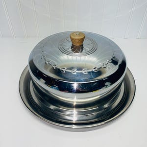 May include: A shiny, stainless steel covered serving dish with a wooden knob on top. The lid has an etched floral design around the edge and a circular pattern in the center. The dish sits on a matching stainless steel base.