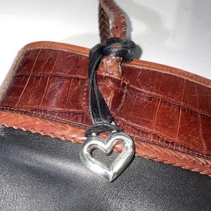 May include: Close-up of a brown and black leather handbag with a silver heart-shaped charm. The bag features a braided strap and a textured, crocodile-like pattern on the leather.
