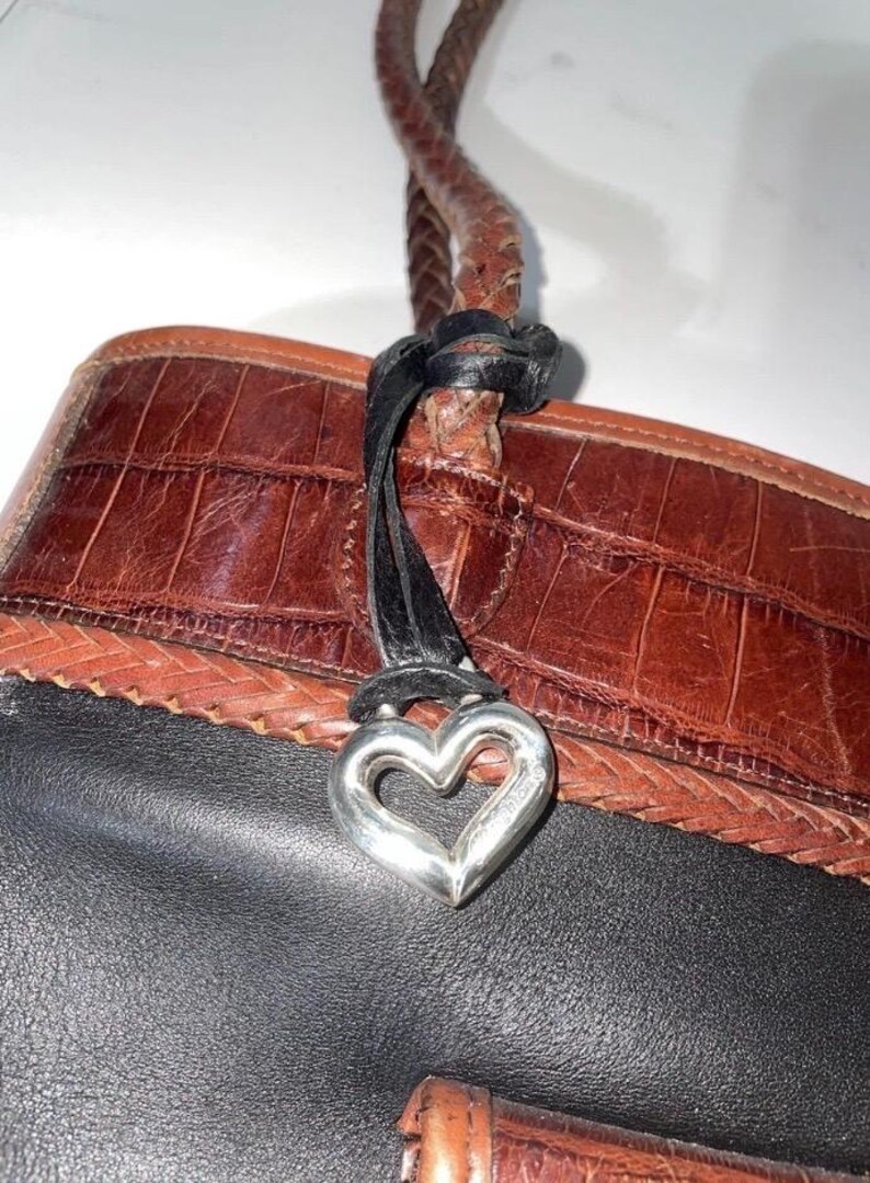 May include: Close-up of a brown and black leather handbag with a silver heart-shaped charm. The bag features a braided strap and a textured, crocodile-like pattern on the leather.