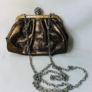 Pode incluir: Uma clutch de cor bronze com uma al&ccedil;a de corrente prateada. A bolsa tem uma apar&ecirc;ncia texturizada e enrugada e um fecho decorativo prateado. A corrente &eacute; drapeada em um la&ccedil;o.