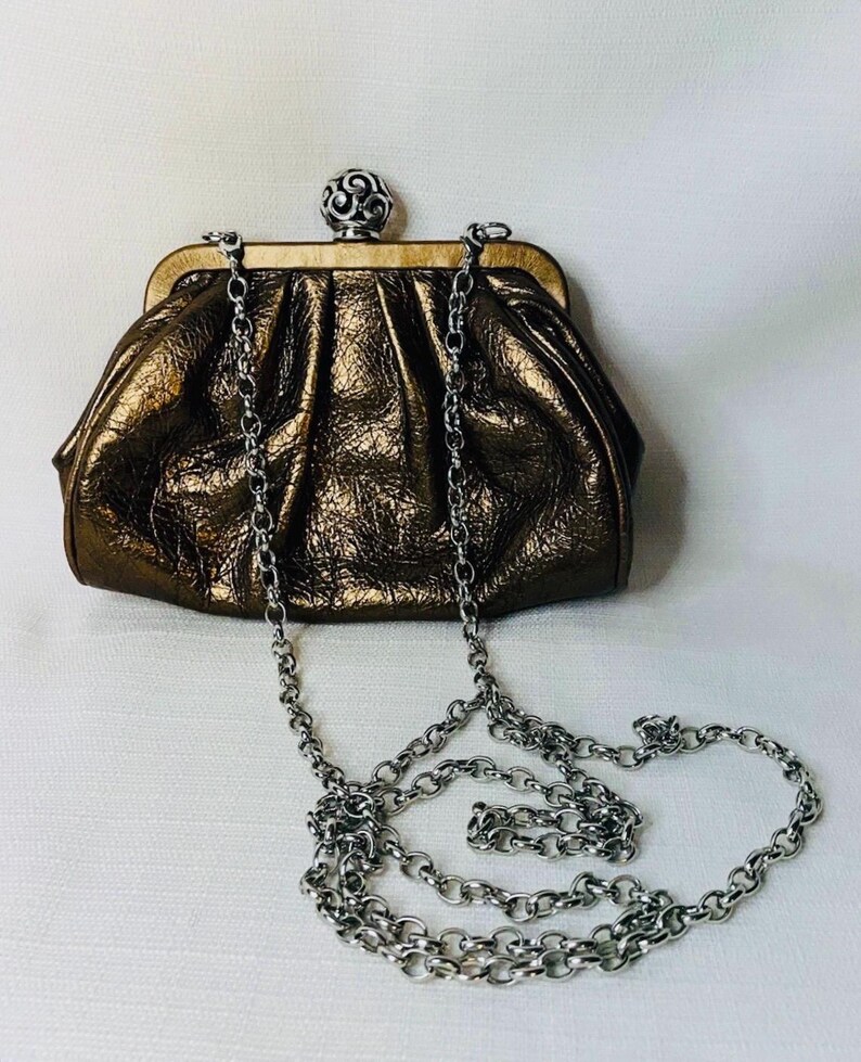 Pode incluir: Uma clutch de cor bronze com uma al&ccedil;a de corrente prateada. A bolsa tem uma apar&ecirc;ncia texturizada e enrugada e um fecho decorativo prateado. A corrente &eacute; drapeada em um la&ccedil;o.