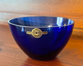 May include: A deep blue glass bowl with a rounded shape. The bowl has a gold label that reads "ALVESTA GLASBRUK SWEDEN". The bowl is sitting on a wooden surface.