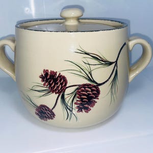 May include: Cream-colored ceramic sugar bowl with a lid and two handles. The bowl is decorated with a pine cone and branch design in shades of brown, green, and black. The rim of the bowl has a dark blue trim.