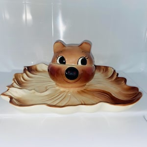 May include: A vintage ceramic bear-shaped trinket dish with a leaf-shaped base. The bear has a brown head with black eyes and a black nose. The dish is beige and brown, with a glossy finish. A charming decorative piece.
