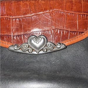 May include: Close-up of a brown leather wallet with a crocodile texture. The wallet features a silver heart-shaped clasp with ornate scrollwork. The background is black.