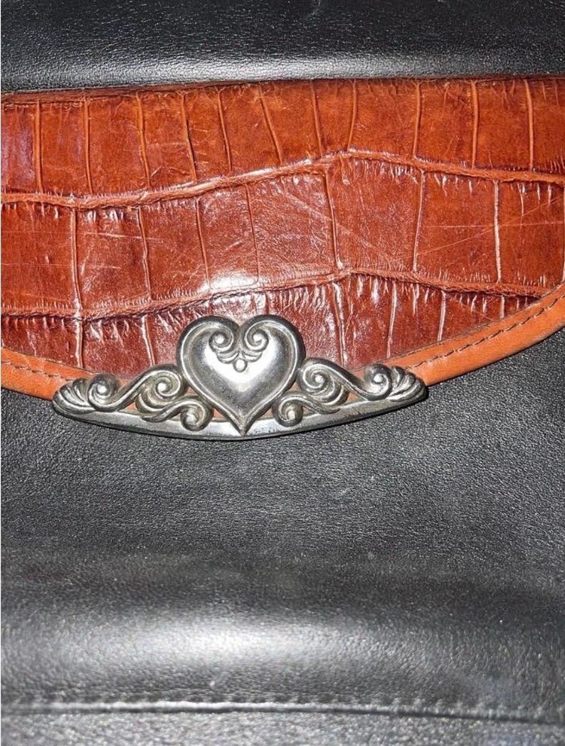 May include: Close-up of a brown leather wallet with a crocodile texture. The wallet features a silver heart-shaped clasp with ornate scrollwork. The background is black.