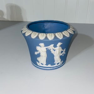 May include: A blue ceramic planter with white relief figures and leaf-like trim. The planter features a flared shape and a detailed design of classical figures. The blue color provides a striking contrast to the white relief.